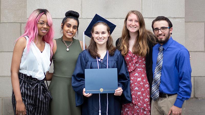 4 students stand with fellow student that's wearing a cap and gown at graduation