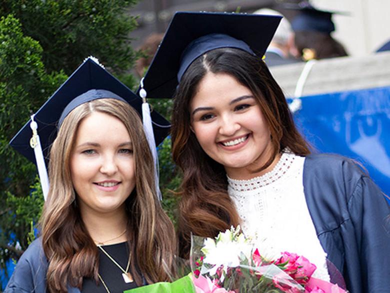 两个面带微笑的女生，头戴毕业帽，身穿毕业礼服，手捧鲜花