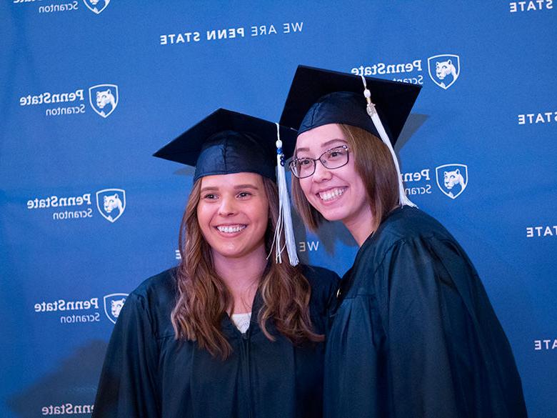 two students in caps and gowns pose in front of Penn State backdrop